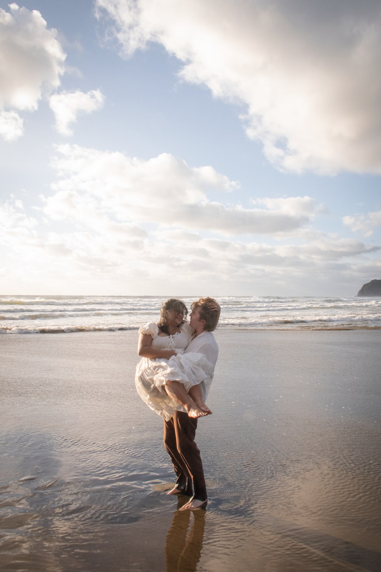 Couple laughing as one carries the other across a wide beach under bright sky