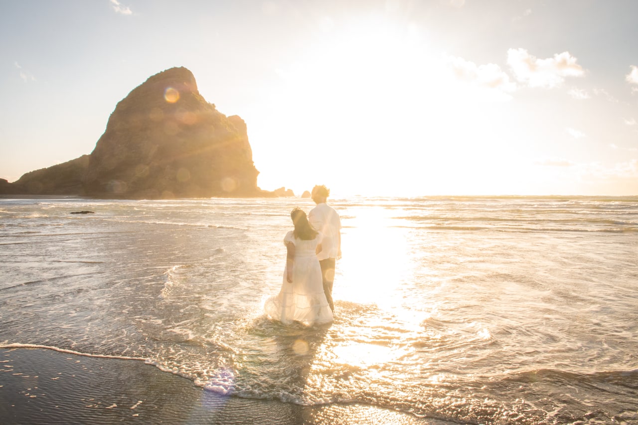 Couple walking into the surf at sunset beside a dark coastal rock formation