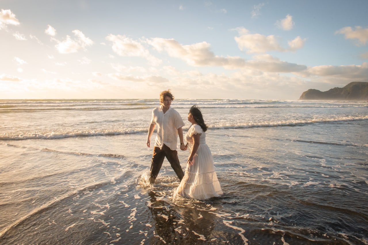 Couple holding hands in shallow surf on a dark-sand beach at sunset