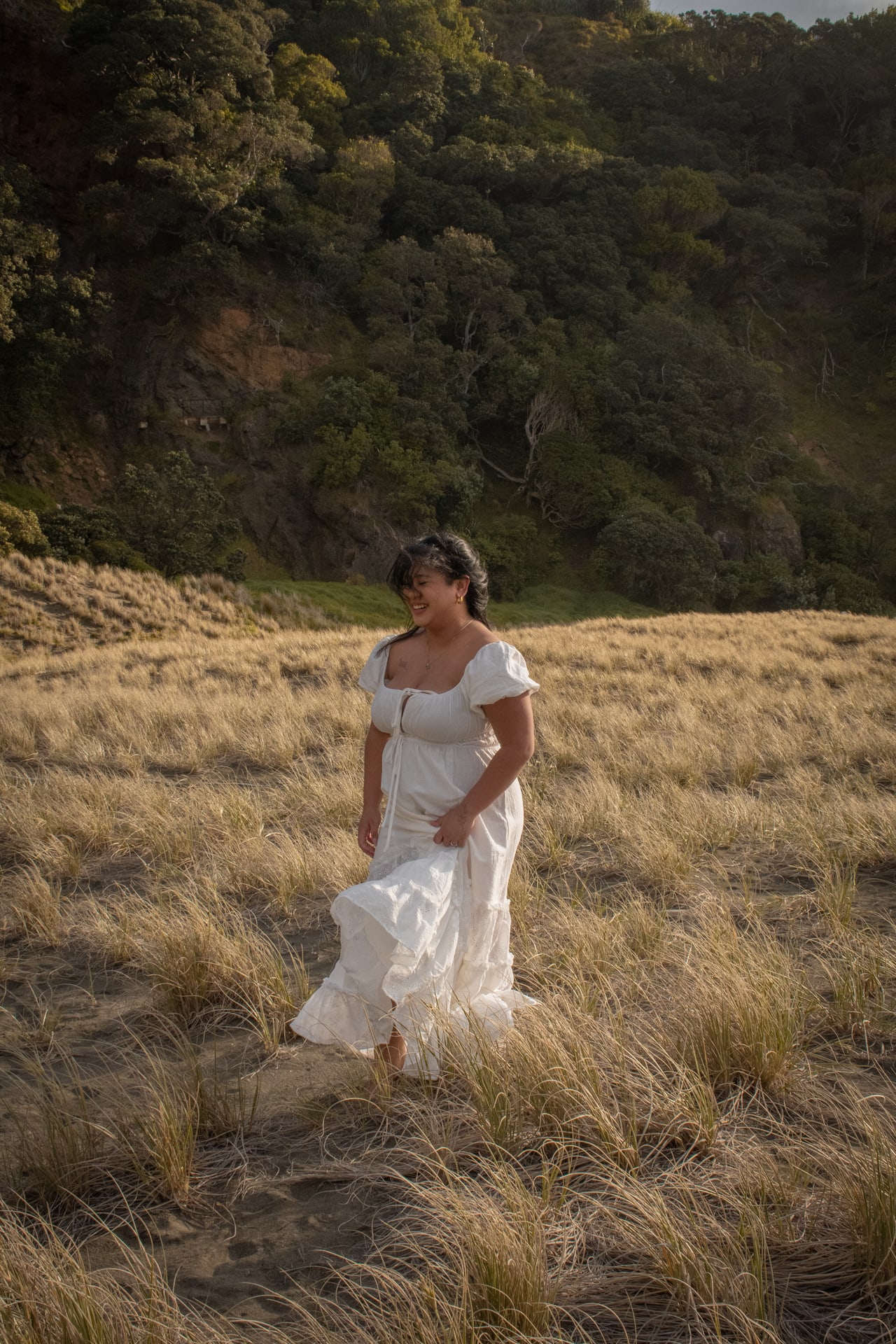 Person in a white dress walking through tall golden coastal grass with a forested hillside behind