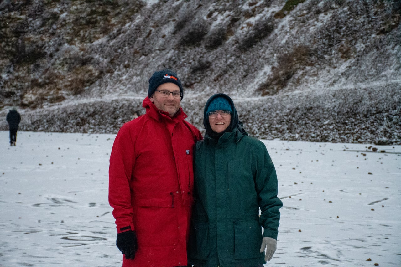Two people in colourful winter coats standing together on snow with a rocky slope behind them