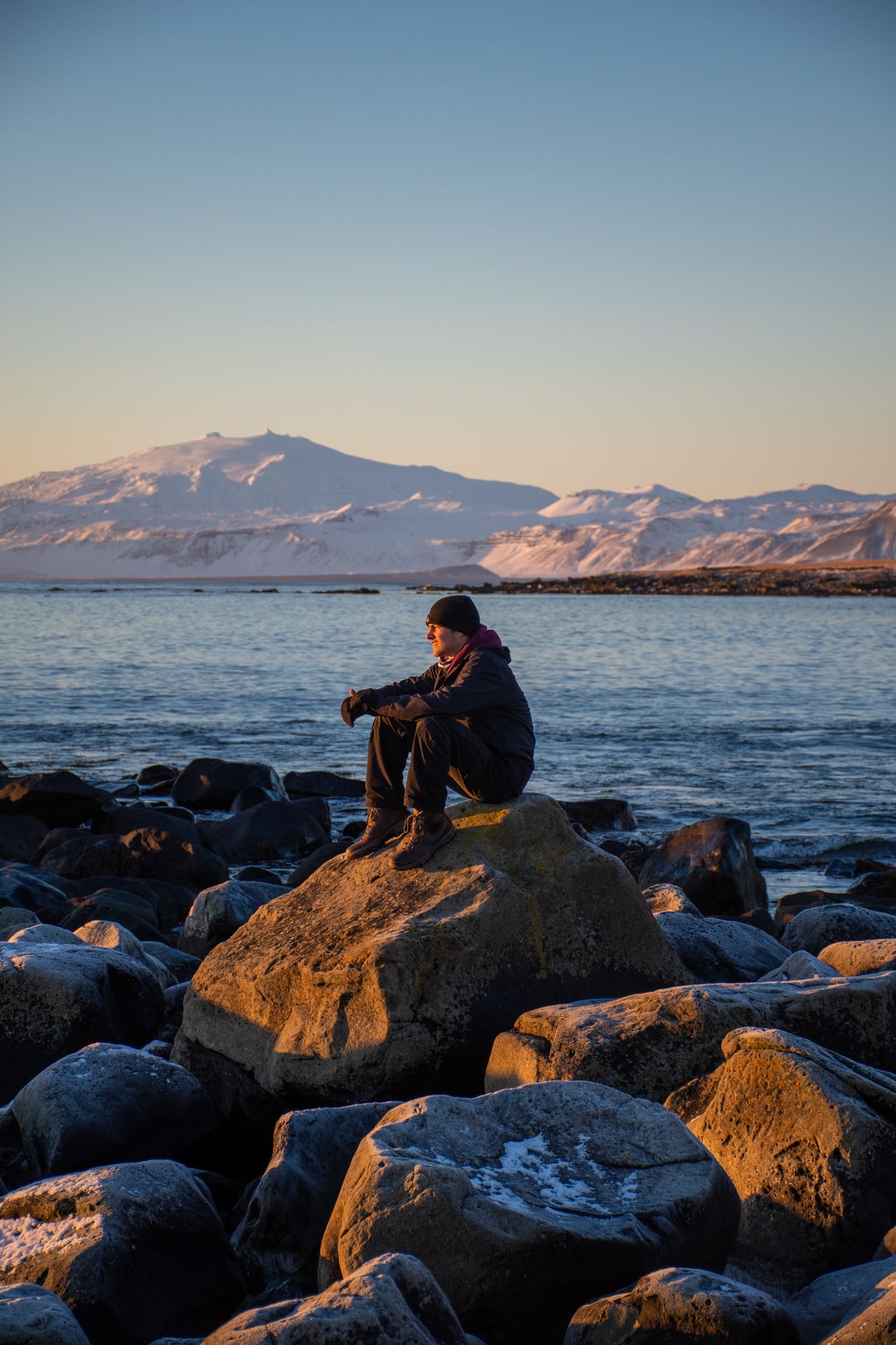 Person seated on shoreline rocks looking toward distant snow-covered mountains at golden hour