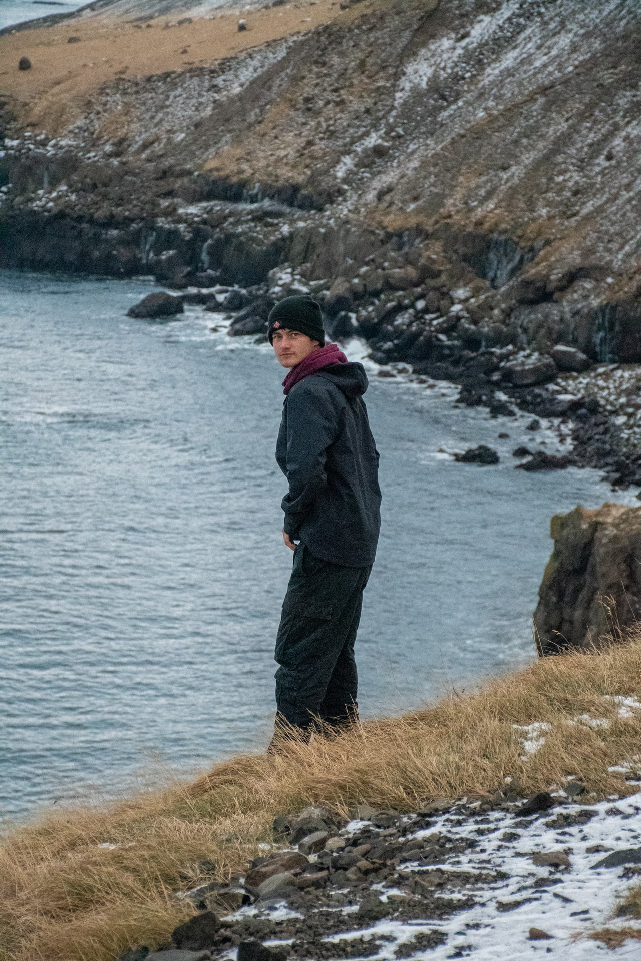 Person in dark outdoor clothing looking back over their shoulder on a grassy coastal cliff above rocky water